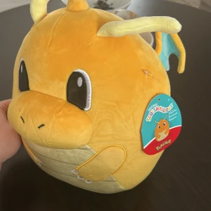 Pokémon Squishmallows Dragonite gosedjur - Supermjuk och kramvänlig Dragonite från Pokémon-serien, tillverkad av plyschmaterial. Gosedjuret är gult med stora svarta ögon, små vingar i blått och gult samt detaljerade klor. Perfekt för Pokémon-fans och samlare.