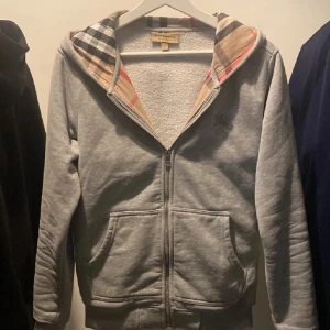 Grå hoodie med dragkedja från Burberry - Säljer en grå hoodie från Burberry med klassiskt rutigt foder i huvan och broderad logga på bröstet. Tröjan har dragkedja framtill, två fickor och ribbade muddar. Perfekt för en avslappnad och stilren look.