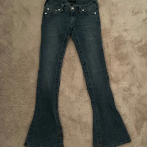 Blå low waisted bootcut jeans från True Religion - Säljer ett par klassiska blå jeans från True Religion med low waisted bootcut passform. Jeansen har fem fickor, silverfärgade knappar och detaljerad söm på bakfickorna. Perfekta för dig som gillar en tidlös jeansstil.