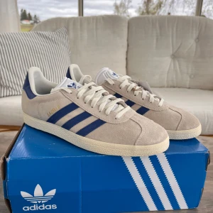 Adidas Gazelle Manchester United Edition - Säljer ett par helt oanvända Adidas Gazelle i beige mocka med blå detaljer och Manchester United-logga på hälen. Skorna har klassiska blå ränder på sidan, vit sula och snörning. Perfekta för dig som vill sticka ut med stilrena sneakers och visa ditt lagstöd.