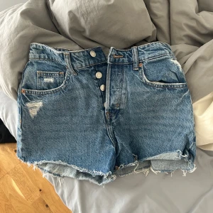Blå jeansshorts med fransar och knappar - Säljer ett par klassiska blå jeansshorts med hög midja, råa fransar nertill och slitningar framtill. Perfekta för sommaren!