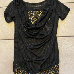 Svart topp  med guldiga detaljer - Svart topp med kort ärm och djup v-ringning. Klänningen har ett nätliknande tyg och dekorativa guldiga fyrkanter framtill och nedtill. Snygg drapering vid bysten och en tight passform. Perfekt för dig som vill sticka ut med coola detaljer.