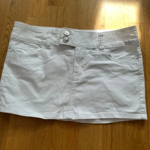 Vit lågmidjad jeanskjol med fickor - Snygg vit kort jeanskjol med två knappar framtill och klassiska fickor både fram och bak. Kjolen har bälteshällor och är perfekt för en fräsch och stilren look. Enkel att matcha med olika toppar.
