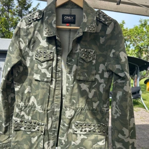Camo jacka med nitar från ONLY - Säljer en grön camouflagejacka från ONLY med coola nitar på bröstfickor, axlar och framtill. Jackan har lång ärm, klassisk krage och flera fickor. Perfekt för dig som har Stockholmsstilen.