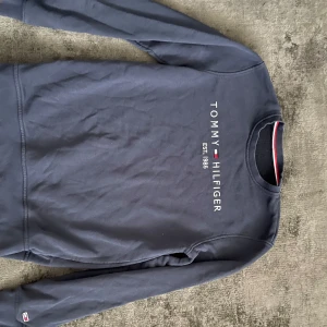 Mörkblå sweatshirt från Tommy Hilfiger - Säljer en mörkblå sweatshirt från Tommy Hilfiger med tryckt logga och texten 'EST. 1985' på bröstet. Tröjan har rund halsringning och långa ärmar. Klassisk och stilren design med små Hilfiger-detaljer vid ärmslut.