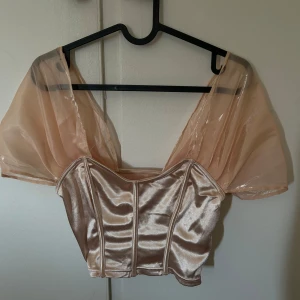 Champagnefärgad satintopp med puffärmar - Snygg croppad topp i glansigt champagnefärgat satinmaterial med transparenta puffärmar. Toppen har en korsettliknande form och markerade sömmar framtill. Dragkedja på sidan. Helt ok stretch men ändå tajt. 