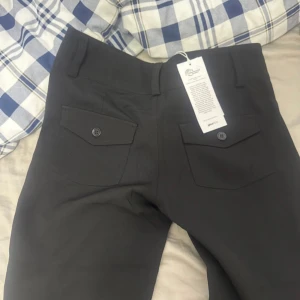 Svarta bootcut kostymbyxor med fickor  - Säljer ett par svarta kostymbyxor med bootcut-passform från Gina tricot som köptes för 499kr. Byxorna har två bakfickor med lock och knapp, samt hög midja och stängs med knappar framtill. Perfekta för en stilren look och passar till många olika outfits. Byxorna är helt nya med prislapp 