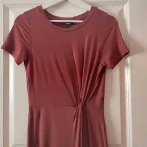 Rostfärgad maxiklänning med knutdetalj - Säljer en rostfärgad klänning i mjukt material med korta ärmar och rund halsringning. Klänningen har en snygg knutdetalj i midjan som ger en fin siluett. Perfekt för dig som gillar enkel men stilren design. Aldrig använd.
