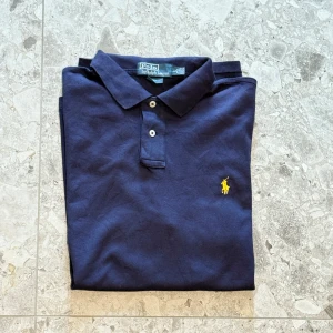 Mörkblå pikétröja från Ralph Lauren - Klassisk mörkblå pikétröja från Ralph Lauren med gul broderad logga på bröstet. Tröjan har korta ärmar, krage och två knappar framtill. Tillverkad i bomull och passar perfekt till en avslappnad stil.