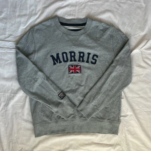 Grå sweatshirt från Morris - Säljer en grå sweatshirt från Morris med mörkblå text och en broderad brittisk flagga på bröstet. Tröjan har rund halsringning och långa ärmar. Klassisk och stilren design med ribbade muddar vid ärmslut och nederkant.