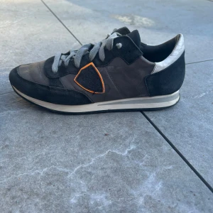 Philippe Model sneakers  - Säljer ett par grå Philippe Model sneakers med svarta mockadetaljer och grå snörning. Skorna har en vit och svart sula samt en orange detalj på sidan. Tillverkade i Italien och har en klassisk, sportig look.
