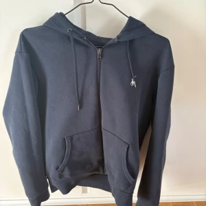 Mörkblå hoodie från Polo Ralph Lauren - Säljer en mörkblå hoodie från Polo Ralph Lauren med dragkedja och huva. Tröjan har två fickor framtill och en liten broderad logga på bröstet. Perfekt för en avslappnad stil.