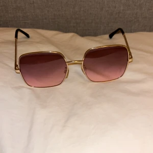 Guldiga fyrkantiga solglasögon med rosa tonade glas - Snygga solglasögon med fyrkantiga linser och tunn guldfärgad metallbåge. Glasen är rosa tonade och skalmarna har en kedjedetalj i guld. Perfekt accessoar för att lyfta din stil.