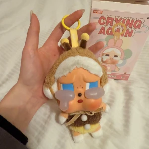 Pop Mart Crying Again nyckelringsväska - Supergullig nyckelringsväska från Pop Mart Crying Again-serien. Väskan är formad som en liten figur med bruna och vita detaljer, mjuk plysch och ett gult nyckelringsfäste. Perfekt som accessoar till väskan eller nycklarna.