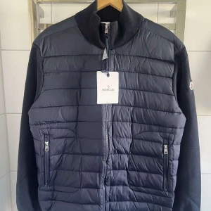 Blå pufferjacka från Moncler - Snygg blå pufferjacka från Moncler med quiltad front och ärmar i stickad kvalitet. Jackan har dragkedja framtill, två fickor med dragkedja och Moncler-logga på ärmen. Perfekt för kyliga dagar. Oanvänd och kommer med kvitto.