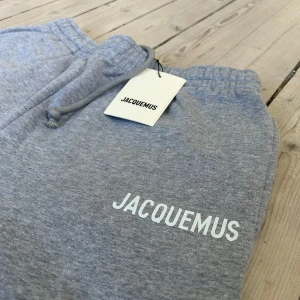 Grå mjukisbyxor från Jacquemus - Snygga grå mjukisbyxor från Jacquemus med vit logga på ena benet. Byxorna har resår i midjan, dragsko och muddar vid bensluten. Perfekta för en avslappnad stil och riktigt sköna att ha på sig.