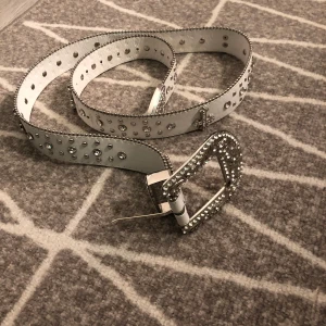 Vitt bälte med nitar och glittriga detaljer - Säljer ett vitt bälte med silvriga nitar, glittriga stenar och silvriga kors, om du gillar lite streetwear/y2k är detta perfekt! 💕jag gjorde små nya hål om bältet är för stort som jag tog bild på.