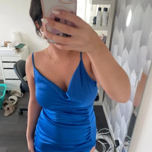 Blå satin klänning - Säljer en snygg blå satin miniklänning från Naanaa köpt från ASOS med smala axelband och draperad detalj framtill. Klänningen har en figurnära passform och dragkedja. Använd 1 gång.