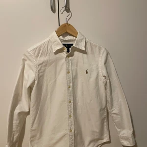 Vit skjorta från Ralph Lauren 12 year - Klassisk vit skjorta från Ralph Lauren med broderad logga på bröstet. Skjortan har lång ärm, knappar framtill och en stilren krage. Perfekt för en clean och tidlös look. 