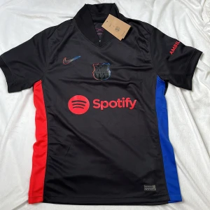 Helt ny FC Barcelona svart fotbollströja Nike Lamine Yamal 19 - Säljer en svart FC Barcelona fotbollströja från Nike med Lamine Yamal 19 på ryggen. Tröjan har röda och blå sidopaneler, klubbmärke och Nike-logga på bröstet samt Spotify som sponsor. Materialet är lätt och andas, perfekt för match eller träning.