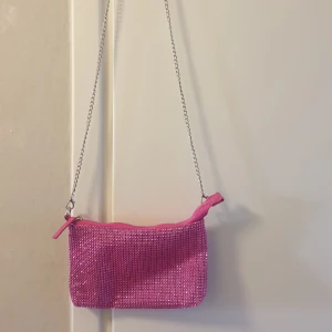 Rosa glittrig axelväska från Primark - Säljer en rosa glittrig axelväska från Primark med silverfärgad kedja. Väskan har dragkedja upptill och är täckt av små glittrande stenar. Perfekt för att addera lite extra bling till din outfit!