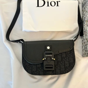 Christian Dior Saddle bag - Säljer en svart axelväska från Christian Dior med justerbart axelband och ikoniskt Dior-mönster. Väskan har lock i skinn, detaljer i metall och ett snyggt spänne framtill med logotyp. Perfekt för dig som vill ha en stilren och lyxig accessoar.
