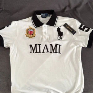 Vit piké MIAMI från Polo Ralph Lauren - Säljer en vit pikétröja från Polo Ralph Lauren med svart broderad logga, MIAMI-text och stora siffror 5 på ärmar och rygg. Pikén har svart krage och ärmar med vita detaljer. 100% bomull och klassisk custom fit. Har inte haft på mig den eftersom den krympte från första gången jag tog på mig den, skulle säga att den passar för en person som är 170-178cm