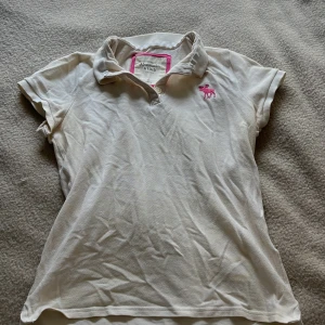 Vit pikétröja från Abercrombie & Fitch - Säljer en klassisk vit pikétröja från Abercrombie & Fitch med rosa broderad älg på bröstet. Tröjan har korta ärmar, krage och knäppning framtill. 