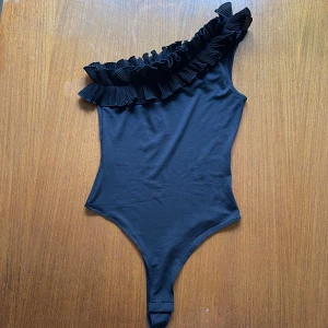 Svart oneshoulder bodysuit med volang - Säljer en svart bodysuit från H&M med en volangdetalj längs ena axeln. Modellen är en oneshoulder och har en tight passform. ALDRIG ANVÄND 