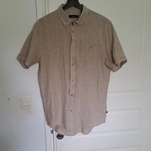 Beige linneskjorta från Matinique - Säljer en beige kortärmad skjorta i linne från Matinique. Skjortan har klassisk krage, knäppning framtill och en bröstficka med knapp. Perfekt för varma dagar och enkel att styla till olika outfits.