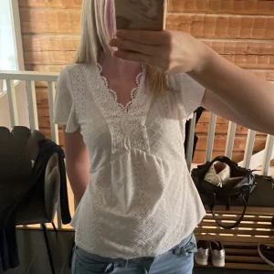 Vit blus med spetsdetaljer i storlek S - Säljer en söt vit blus med v-ringning och spetsdetaljer längs halsringningen och bröstet. Blusen har korta ärmar och ett broderat mönster över hela tyget. Perfekt till jeans eller kjol!
