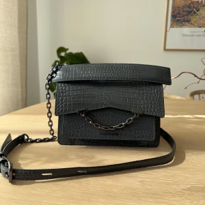 Svart axelväska i krokopräglat skinn från Karl Lagerfeld - Säljer en svart axelväska från Karl Lagerfeld i krokopräglat skinn. Väskan har en kedjedetalj framtill, magnetlås och justerbar axelrem. Perfekt storlek för det viktigaste och stilren design med exklusiv känsla.