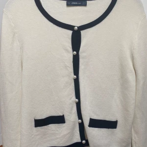 Kofta från Zara - Säljer en stilren vit kofta från Zara med svarta kanter och dekorativa pärlknappar. Perfekt för en klassisk och chic look.