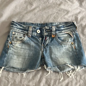 Blå low waist jeansshorts - Säljer ett par blå low waist jeansshorts från Replay med snygga fransade kanter och slitningar. Shortsen har snygga bakfickor och sitter supersnyggt. De har en dekorerad patch bak med nitar och paljetter. Perfekta för sommaren!🥰 Skriv vid frågor, går att diskutera priset😊