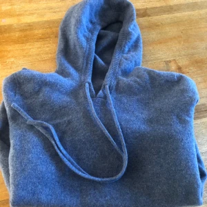 Blå hoodie i kashmir - Mysig blå hoodie i mjuk kashmir med huva och dragsnören. Perfekt för en avslappnad och stilren look. Ribbstickade muddar vid ärmslut och nederkant.