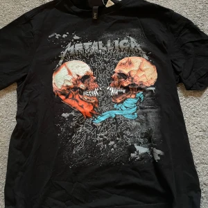 Svart Metallica t-shirt från H&M - Svart t-shirt från H&M med Metallica-tryck framtill. Motivet visar två dödskallar i färg med detaljer i rött och blått. Regular fit och kortärmad modell, perfekt för dig som gillar bandtröjor och rockstil.