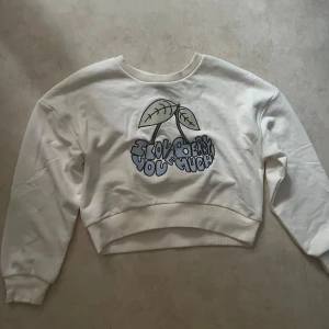 Vit sweatshirt med körsbärstryck från LMTD - Vit sweatshirt från LMTD med ett stort tryck av körsbär och texten 'I love you cherry much' i blått framtill. Tröjan har rund halsringning, långa ärmar och ribbade muddar vid ärmslut och nederkant. Aldrig använt.