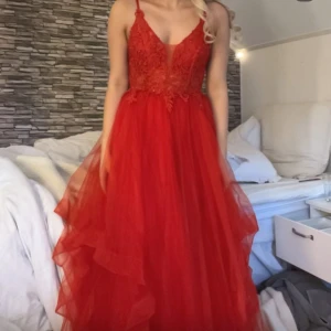 Röd långklänning med spets och volang - Säljer en röd långklänning med smala axelband, v-ringning och vacker spets upptill. Kjolen är i flera lager tyll med volangdetaljer för extra volym. Perfekt för bal eller fest. Klänningen har en romantisk och elegant känsla.