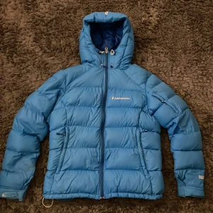 Blå dunjacka med huva från Peak Performance - Säljer en blå dunjacka från Peak Performance i dammodell, storlek L. Jackan har huva, dragkedja framtill och två fickor med dragkedja. Perfekt för kyliga dagar och håller dig varm med sin pufferdesign.