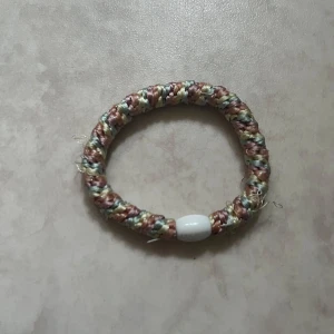 Flätat armband med pärldetalj - Säljer ett handgjort armband i flätad textil med beige, gröna och bruna toner. Armbandet har en vit pärldetalj som accent. Perfekt accessoar för en personlig stil.