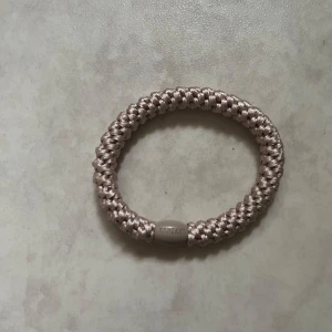 Beige flätat armband från kknekki - Säljer ett beige armband från kknekki med flätad design i glansigt syntetmaterial. Armbandet har en oval pärldetalj med märkesnamnet ingraverat. Perfekt accessoar för att lyfta din stil.