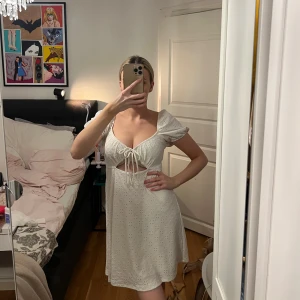 Vit klänning - Söt vit kortklänning med broderade hål, puffärmar och knyt framtill. Klänningen har en v-ringad urringning och öppen detalj under bysten. Perfekt för sommarens alla tillfällen. 