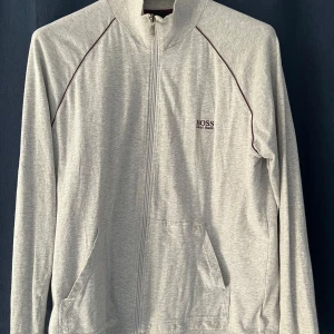 Ljusgrå zip-jacka från Hugo Boss - Säljer en ljusgrå långärmad zip-jacka från Hugo Boss med diskret logga på bröstet. Jackan har dragkedja framtill, två fickor och tunna mörka linjer längs axlarna. Perfekt för en stilren och avslappnad look.