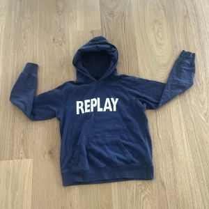 Blå hoodie från Replay - Säljer en blå hoodie från Replay med stor vit logga på bröstet. Tröjan har huva och en klassisk magficka framtill. Perfekt för en avslappnad och cool stil.