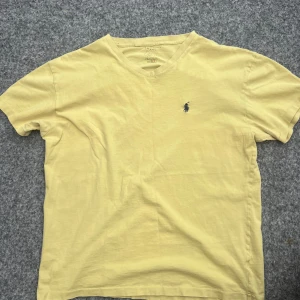 Gul t-shirt från Polo Ralph Lauren - Säljer en ljusgul t-shirt från Polo Ralph Lauren med klassisk blå broderad logga på bröstet. T-shirten har rund halsringning och korta ärmar. Perfekt för en avslappnad och stilren look.