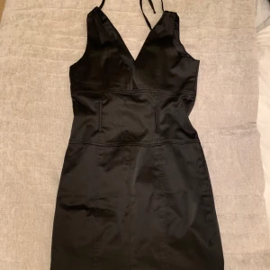 Svart kortklänning med v-ringning från Loleumary - Säljer en svart kortklänning från Loleumary med smala axelband och djup v-ringning. Klänningen har figurnära passform och är tillverkad i ett slätt, glansigt material. Perfekt för dig som gillar en stilren och enkel look.
