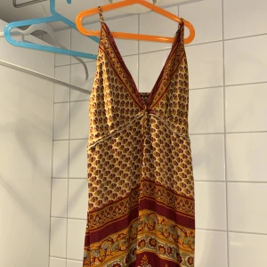 Mönstrad långklänning från Shakaloha - Säljer en bohemisk långklänning från Shakaloha med smala axelband och v-ringning. Klänningen har ett detaljerat mönster i vinrött, gult och beige med paisley och geometriska inslag. Perfekt för sommarens varma dagar.