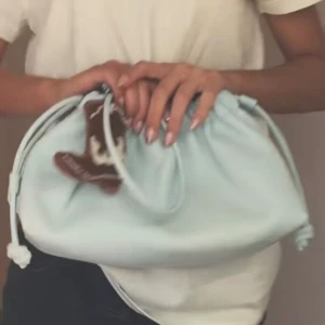 Ljusblå axelväska med dragsko - Säljer en ljusblå axelväska med dragskostängning och rundad form. Väskan har en mjuk look och är perfekt för att bära det viktigaste. En liten brun accessoar hänger på sidan som extra detalj.