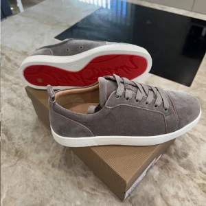 Gråa sneakers från Christian Louboutin - Säljer ett par gråa sneakers från Christian Louboutin med vit sula och röd undersida. Skorna har snörning och är tillverkade i mocka. Kommer med originalkartong och dustbag. Dom är provad men nästan helt nya
