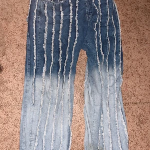 Blå jeans med slitna ränder från Review - Unika blå jeans från Review med baggy fit och tydliga slitna vertikala ränder över hela byxan. Jeansen har klassisk femficksdesign och knappgylf. Perfekt för dig som vill sticka ut med en cool streetstil.